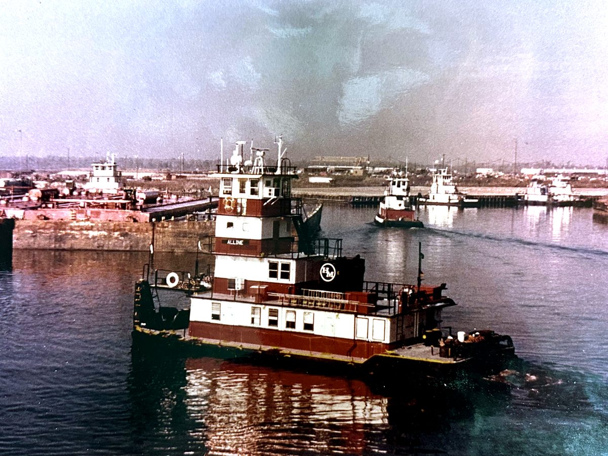 The towboat M/V Alline underway in Hollywood Marine colors, with towboats and a dock along the bank behind her