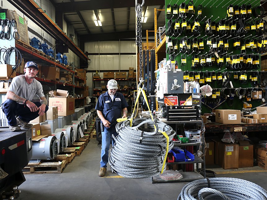 Two of the parts crew in the warehouse, a coil of wire rope slung from the hoist beside racks of belts, filtration, and drive parts